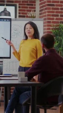 Dikey video: Asyalı kadın toplantı odasında grafik ve grafikleri açıklıyor. Çeşitli meslektaş grupları yönetim projesi için iş araştırmalarını paylaşmak üzere toplanıyorlar. Takım çalışması brifingi.