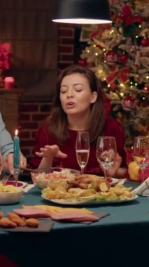 Vertical video: Happy wife talking to guests about food while sitting together at Christmas dinner table. Festive cheerful people enjoying holiday inside cozy aparment with traditional and authentic