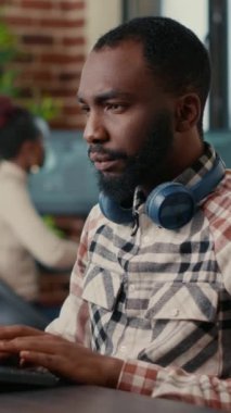 Vertical video Portrait of focused african american programer wearing wireless headphones working looking at computer screen while typing. Software developer writing code in it agency office.
