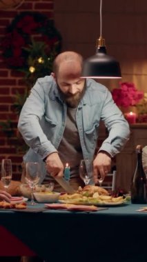 Dikey video: Gururlu koca evde Noel yemeğinin tadını çıkarırken konuklar için kızarmış tavuk kesiyor. Şenlikli adam, Xmas 'ın süslü dairesinde geleneksel yemekle kış tatilinin tadını çıkarıyor.