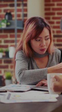 Vertical video: Team of asian women analyzing business charts on laptop and papers in office. Coworkers planning development strategy with financial statistics at briefing meeting. Sales research