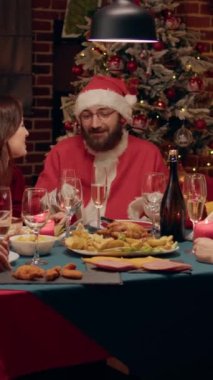Vertical video: Joyful man disguised as Santa Claus clinking champagne glass with family enjoying Christmas dinner. Festive relatives celebrating winter feast while sitting at table with Santa Claus
