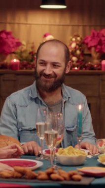 Dikey video: Mutlu adam yaşlı kadınla konuşuyor ve Noel yemeğinin tadını çıkarırken kameraya gülümsüyor. Neşeli aile üyeleri kış tatilini kutlarken evde oturuyorlar. El bilgisayarı