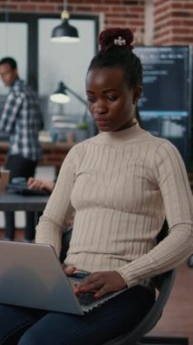 Vertical video Portrait of african american programer sitting down coding on laptop looking up and smiling at camera. Coder using portable computer next to mixed team in software development agency.