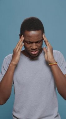 Dikey video: Üzgün endişeli adam stüdyoda acı veren baş ağrısıyla uğraşıyor, kendini kötü ve bitkin hissediyor. Stresli insan acı çekiyor, hastalık sorunu yaşıyor ve ciddi migreni tedavi etmek için tapınakları ovuyor..