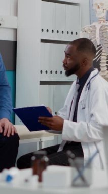 Dikey video: Genel pratisyen hekimin tıp kabinesinde kıdemli hastaya danışması, sağlık kontrolünde hastalık teşhisi hakkında konuşması. Klinikte yaşlı bir adamla sağlık muayenesi yapıyorum.