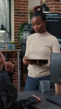 Vertical video Software developer standing up holding digital tablet talking with programer sitting at desk with computer screens showing running code. System engineer coding with team writing machine