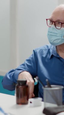 Dikey video: Tekerlekli sandalyede maske takan yaşlı bir hasta tıbbi kabine uzmanı ile sağlık kontrolüne gidiyor. Kronik engelli yaşlı bir adamın asistanıyla sağlık hizmeti randevusu var.