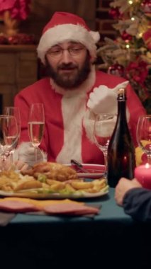 Dikey video: Noel Baba kılığında komik bir adam evde Noel yemeği yerken ailesiyle hediyeler hakkında konuşuyor. Geleneksel kostümlü, kış tatilini birlikte kutlayan neşeli koca.