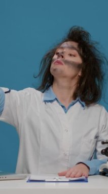 Vertical video: Crazy amusing chemist analyzing lab beaker and acting insane, being foolish and doing goofy playful facial expressions. Mad silly scientist with dirty face and wacky funny hair.