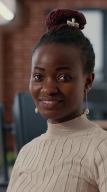 Vertical video Portrait of smiling african american software developer looking at camera in artificial intelligence development agency. Confident programer posing next to colleagues working in big