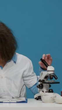 Dikey video: Arka planda laboratuvar mikroskobu kullanan aptal kimyager aptalca davranıyor ve sırıtıyor. Dağınık saçlı, aptal, komik, çılgın, eğlenceli saç stili olan çılgın bilim adamı stüdyoda..