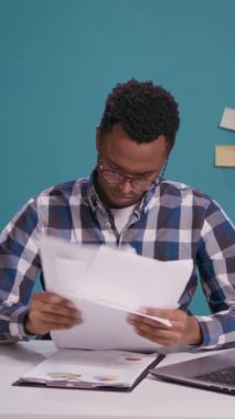 Dikey video: Genç iş adamı kağıtlar üzerinde mali tabloları düzenliyor, dizüstü bilgisayarla istatistik planı oluşturmak için belgeleri analiz ediyor. Pazarlama grafikleriyle iş dosyalarına bakan hayal kırıklığına uğramış kişi.