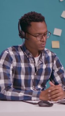 Dikey video: Genç bir adam tele pazarlama yapmak için mikrofonlu bir kulaklık kullanarak yardım hattında müşteriyle konuşuyor. Müşteri hizmetleri çalışanı telesallerden bahsediyor..