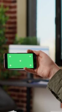 Vertical video: Office employee looking at horizontal greenscreen on smartphone, analyzing isolated copyspace on template. Working with blank chroma key and mockup background on mobile phone. Tripod