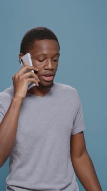 Dikey video: Milenyum adamı mavi arkaplanda telefonla konuşuyor, dokunmatik ekranlı kablosuz telefona cevap veriyor. İnternet bağlantısında uzaktan konuşma yapmak için cep telefonu hattını kullanan kişi.