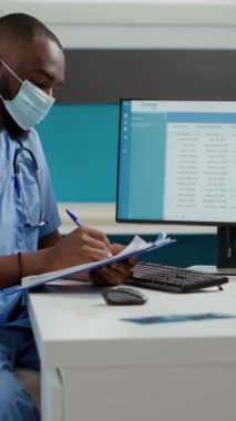 Dikey video: Sağlık çalışanı fiziksel engelli yaşlı kadına danışmanlık yapıyor, klinikte check-up yapıyor. Hemşire randevusunda felçli hasta ve maskeli kocasıyla konuşuyor..