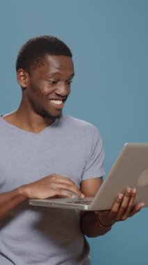 Dikey video: Sosyal medya ve çevrimiçi web sitesinde gezinmek için taşınabilir elektronik bilgisayar kullanan neşeli kişi, stüdyoda dizüstü bilgisayarda internette geziniyor. Elinde kablosuz aygıt tutan mutlu adam..