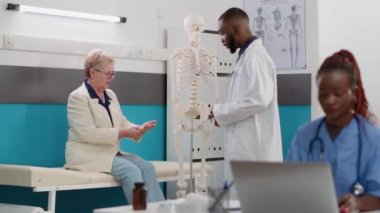 Erkek osteopat, yaşlı kadına insan iskeleti kemiklerini inceliyor, hastaya ortopedi teşhisi koymak için anatomi omuriliğini analiz ediyor. Klinikte Osteopati danışmanlığı randevusu.