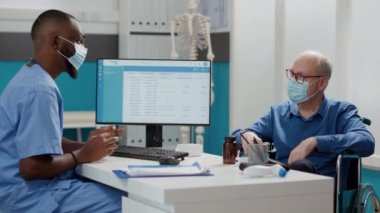 Afrikalı Amerikalı hemşire yaşlı bir adamla istişarede bulunuyor. Tekerlekli sandalyedeki fiziksel bozukluklarla ilgileniyor. Covid 19 salgını sırasında hastaya check-up randevusu almak.