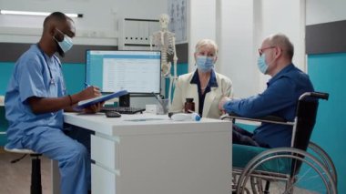 Sağlık kontrolünde fiziksel engelli yaşlı bir adama danışan erkek hemşire yüz maskeli hasta ve eşiyle konuşuyor. Tekerlekli sandalye kullanıcısıyla görüşme, engelli desteği.