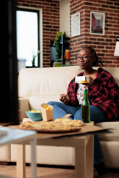 African american woman watching film on television and eating noodles from takeaway food delivery, having fun. Enjoying takeout meal in package box with beer bottles, watch tv movie.