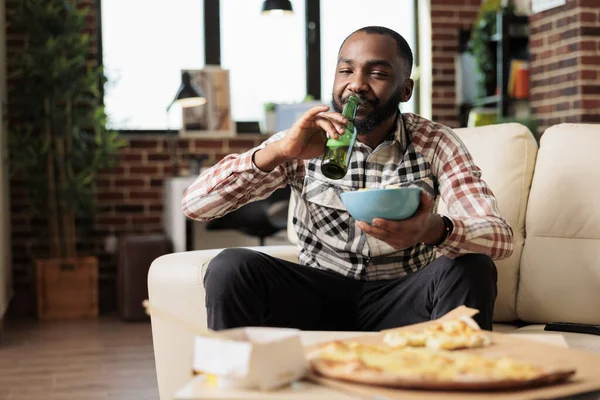 Şişeden alkollü bira içen ve elinde cipslerle televizyon programında film izleyen neşeli bir yetişkin. Paket servis, atıştırmalık, eğlence ve içeceğin tadını çıkarıyorum..