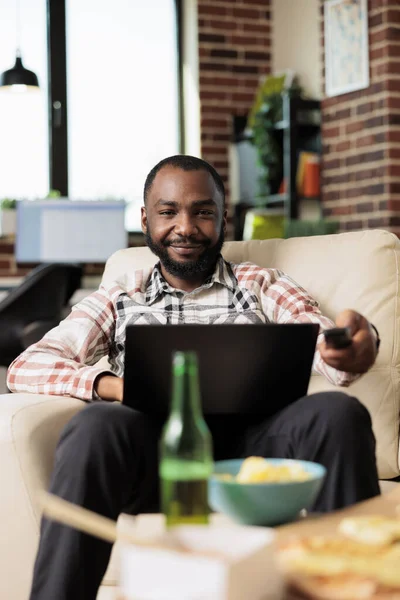 African american man working on laptop and switching tv channels with remote control, browsing internet and finding film to watch. Eating takeout delivery fast food meal at home.