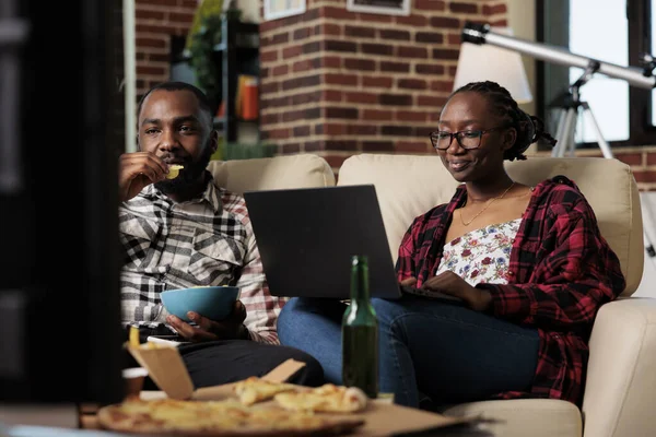 Young couple watching movie on tv and laptop to browse internet, eating fast food takeaway delivery at home. Using computer in front of television, eat pizza and chips with beer bottles.