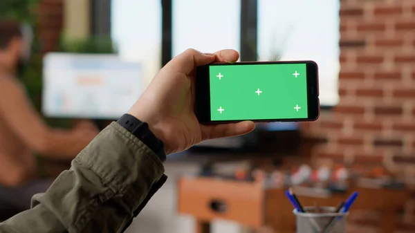 Business manager holding horizontal greenscreen on smartphone, looking at blank chroma key copyspace on display. Using isolated mockup template or background on telephone. Close up.