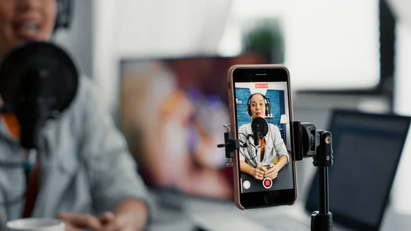 Social media content creator recording vlog with touchscreen phone while talking to audience. Joyful internet star sitting at home studio desk while broadcasting video blog. Close up