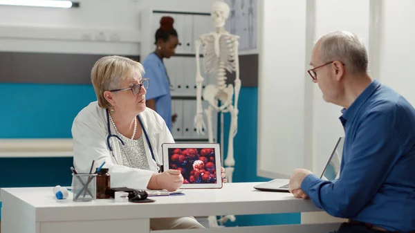 Female physician holding digital tablet with coronavirus animation, explaining pandemic disease to retired person at checkup visit. Virus illustration on device screen, covid 19 prevention.