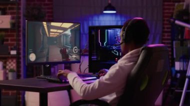African american gamer having intense experience streaming online first person shooter game on pc station at home. Man playing intense action game in tournament talking with team using gaming headset.
