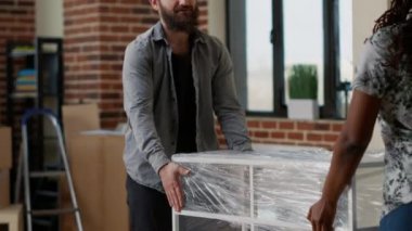 Çeşitli çiftler oturma odasında taşınmak için masa taşıyorlar. Birlikte yeni bir ev eşyası satın aldıktan sonra eşyaları yerleştiriyorlar. Kiralık ev dekorasyonu. El kamerasıyla..