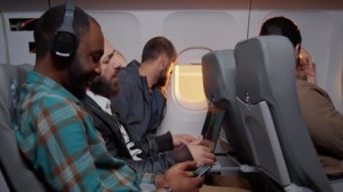 Uçakta ticari havayolu ile uçan çeşitli yolcular, zaman geçirmek için akıllı telefon, dizüstü bilgisayar ve cihazlar kullanıyorlar. Tatil yerine varmak için gün batımında seyahat ediyorlar. El kamerasıyla..
