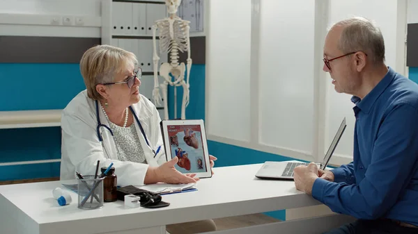 Female physician showing cardiology diagnosis on digital tablet, holding gadget with cardiovascular system illustration to retired man. Heart condition and disease at checkup visit.