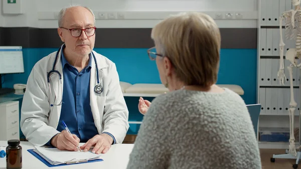 Senior physician taking examination notes on checkup files report, giving advice and medication to retired adult at appointment. Writing disease and diagnosis information on papers.