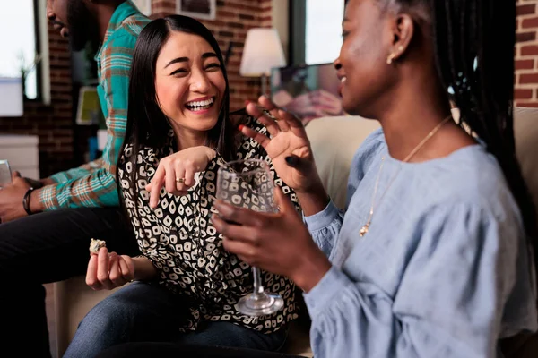 Cheerful smiling diverse women sitting on sofa at wine party while enjoying fun time together. Multiethnic happy people celebrating birthday event while drinking alcoholic beverages.