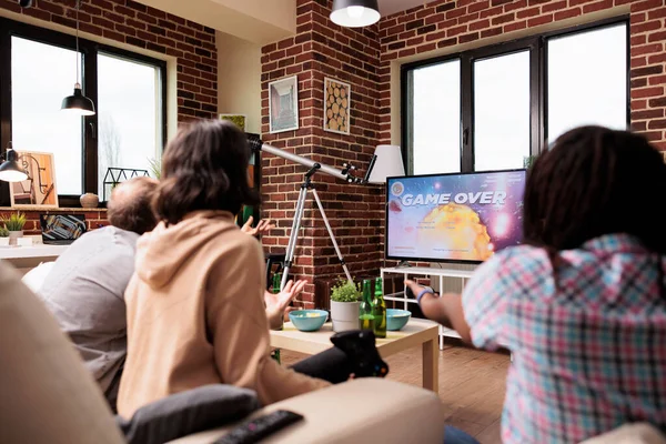 Upset woman reacting to game over screen while losing game round on modern console. Casual game players angry about lost match round while sitting on sofa in living room with close friends group.