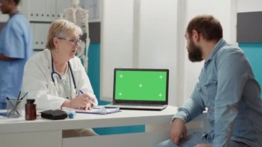 Senior doctor and patient looking at laptop with greenscreen at checkup visit. Man and woman using isolated template with blank chromakey background and mockup copyspace on display.