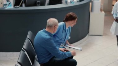 Multiethnic group of patients in waiting area lobby, preparing to attend medical checkup visit with physician. Healthcare examination in professional facility, hospital reception desk. Handheld shot.