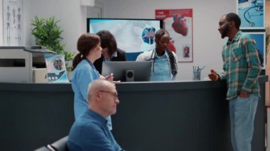 Diverse group of people talking about healthcare at reception counter, medical staff and patient discussing checkup procedure. Waiting room area at hospital lobby for appointment.