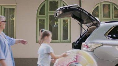 Grandparents taking niece to beach while loading luggage in car trunk. Senior people with granddaughter putting voyage baggage and trolleys inside vehicle while going on summer field trip.