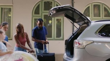 Çok ırklı insanlar yaz tatiline çıkarken bir yandan da bagajlara ve yük arabalarına yükleme yapıyorlar. Aile ve Afrikalı Amerikalı arkadaşlar bagajlarını arabaya koyarken okul gezisine çıkıyorlar..