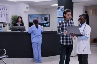 Özel klinikteki Asyalı hastalara laboratuvar sonuçlarını sunan bir dizüstü bilgisayar tutan Afrikalı Amerikalı doktor. Doktor genç adamla konuşuyor. Taşınabilir bilgisayardaki randevuyu kontrol ediyor..
