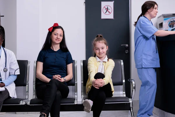 Smiling mother and daughter in busy private clinic waiting for pediatric doctor to do routine checkup while nurse is talking with receptionist. Woman and her child attending pediatrician appointment.