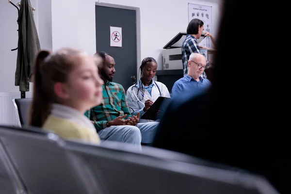 Diverse people waiting in private clinic while hospital medic is asking medical questions in waiting room. Selective focus on african american doctor with stethoscope completing form for patients.