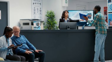 Kadın resepsiyonist, hastalara sağlık hizmetleri randevusu için uzman ile tıbbi rapor doldurmalarında yardımcı oluyor. Hastane resepsiyonunda muayene için bekleme salonu..