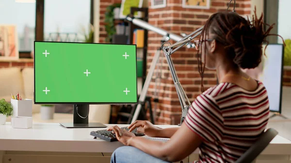 African american freelancer working from home with greenscreen on computer, at desk. Using chroma key template with isolated mockup display and blank copyspace background. Tripod shot.