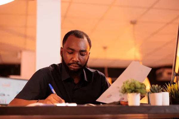 African american employee taking notes about startup contract, analyzing paperwork with business data in office. Working with documents to do research and find executive solutions.